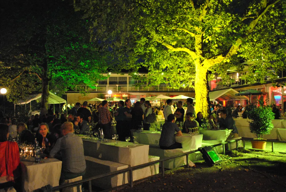 Outdoor evening social event with a large crowd standing and sitting at white banquet tables, illuminated by dramatic green lighting on a large tree and surrounding foliage