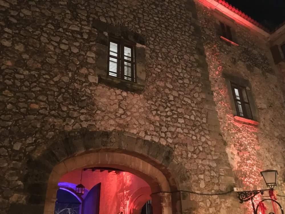 Nighttime view of a rustic stone building facade with an arched entrance and shuttered windows, illuminated by dramatic pink and red event lighting