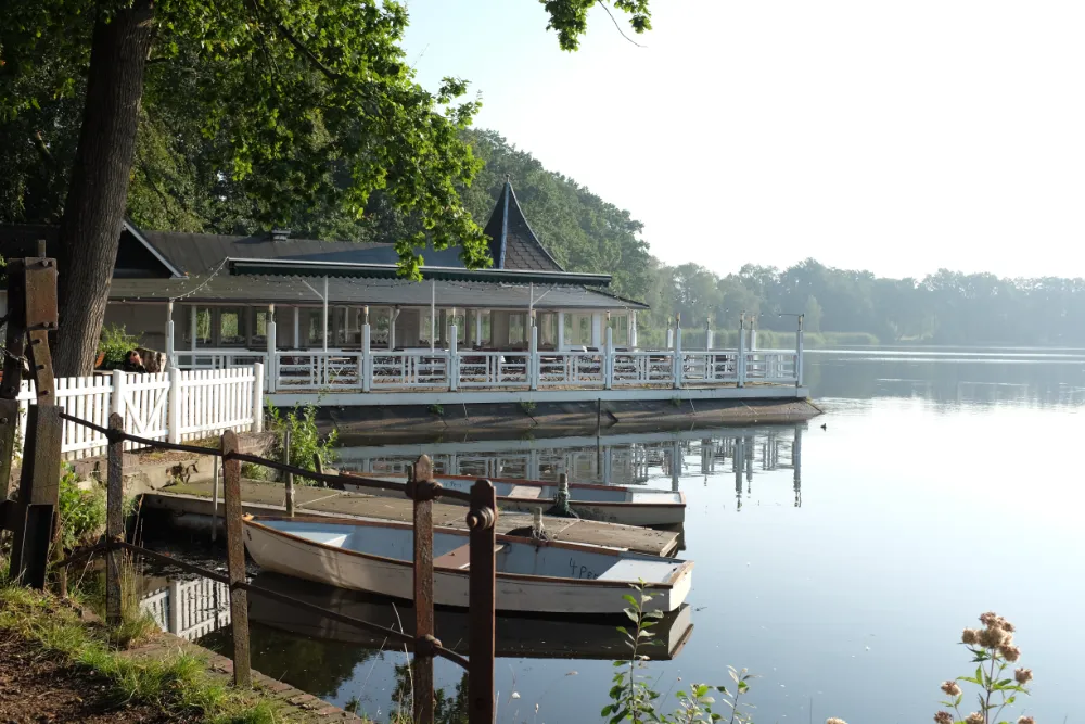 Lakeside restaurant with a white wooden terrace extending over calm water, viewed past a rustic fence and docked rowboats in the foreground under a bright sky