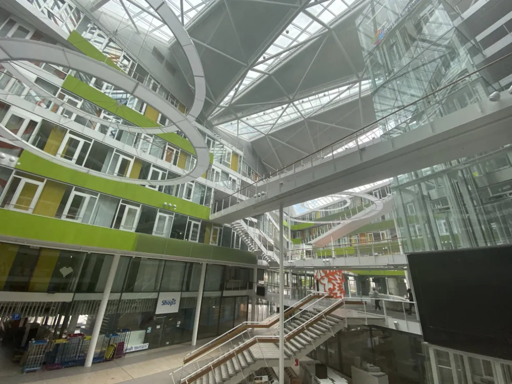 Interior view of a massive, modern multi-story atrium with a glass roof, featuring distinctive lime-green wall panels, crisscrossing white walkways, and a central staircase