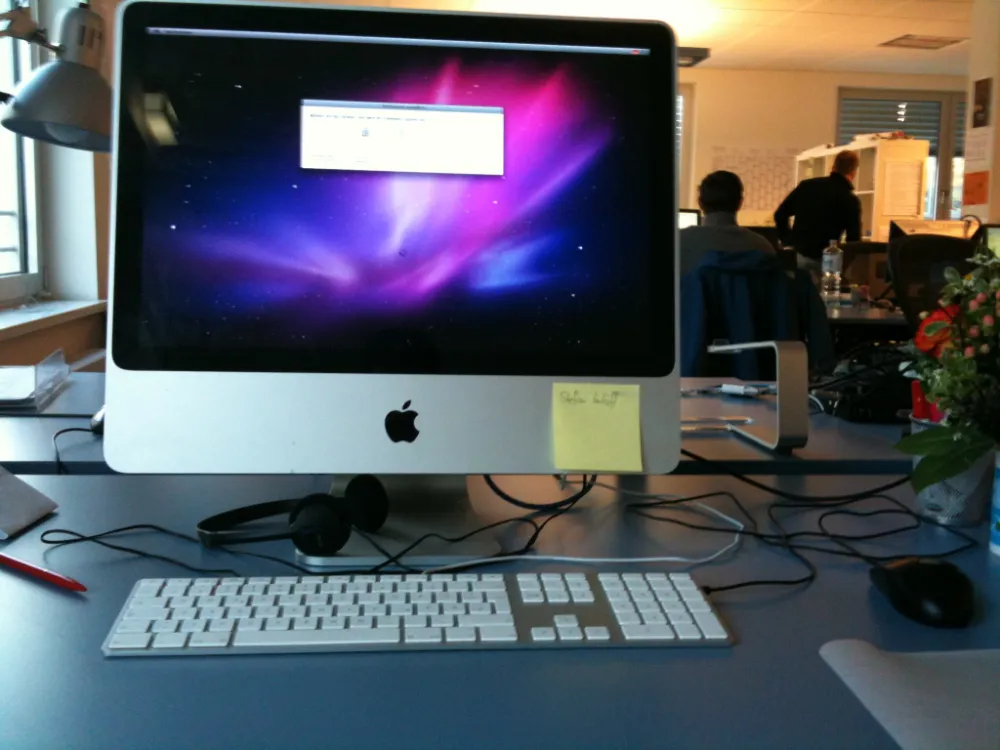 Apple iMac desktop workstation in an office with a wired keyboard, headset, and a yellow sticky note attached to the monitor, with colleagues working in the background