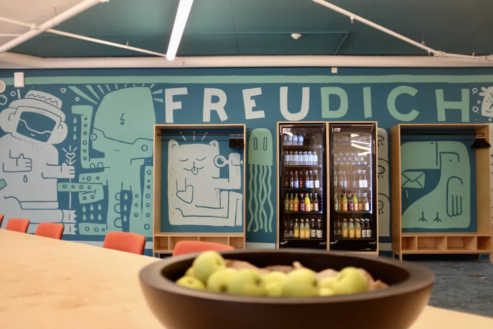Office break room featuring a large teal wall mural with the text 'FREU DICH' and cartoon illustrations, framing beverage refrigerators and wooden coat racks, with a bowl of green apples in the foreground