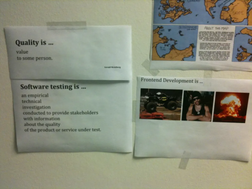 Office wall with taped signs defining 'Quality' and 'Software testing', alongside a humorous poster titled 'Frontend Development is …' featuring images of a monster truck, a man in sunglasses, and a large explosion