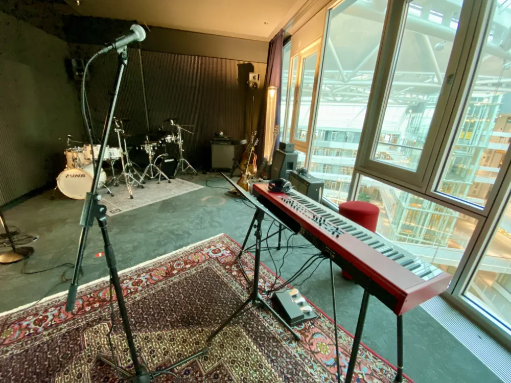 Music rehearsal room with floor-to-ceiling windows overlooking an atrium, equipped with a red keyboard, microphone stand, drum kit, guitars, and amplifiers, set on Persian-style rugs