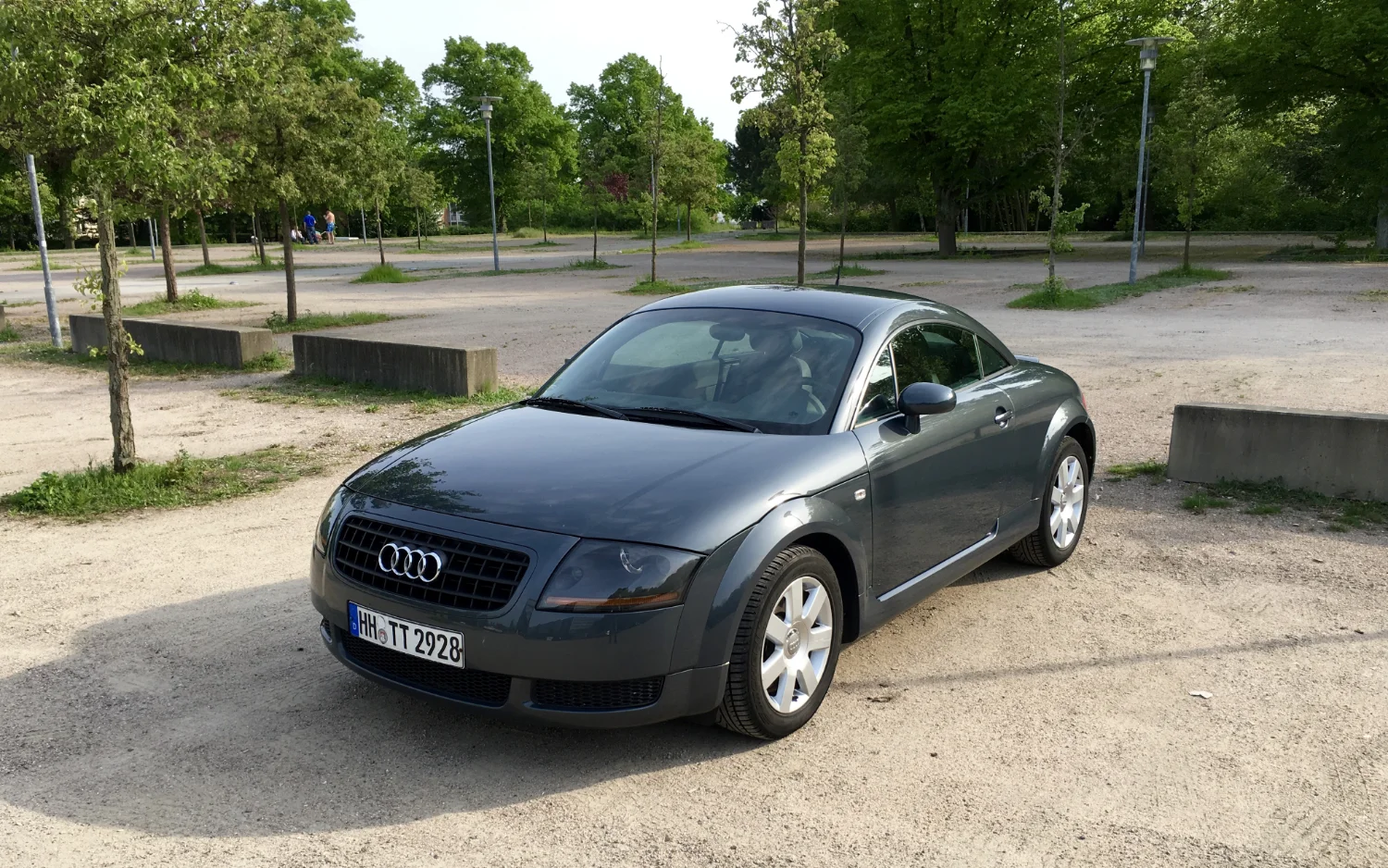 Dolomite-grey Audi TT Coupé parked on a sunlit gravel lot, viewed from the front left with trees behind it.