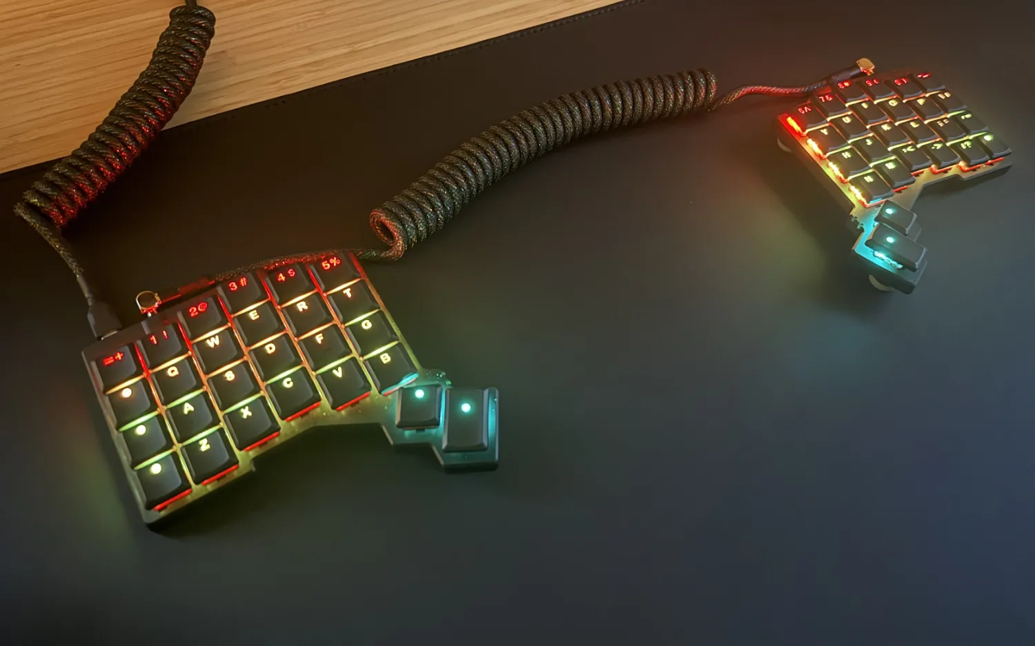A close-up of a split ergonomic keyboard with colorful backlighting, connected by a coiled cable, on a dark desk surface.