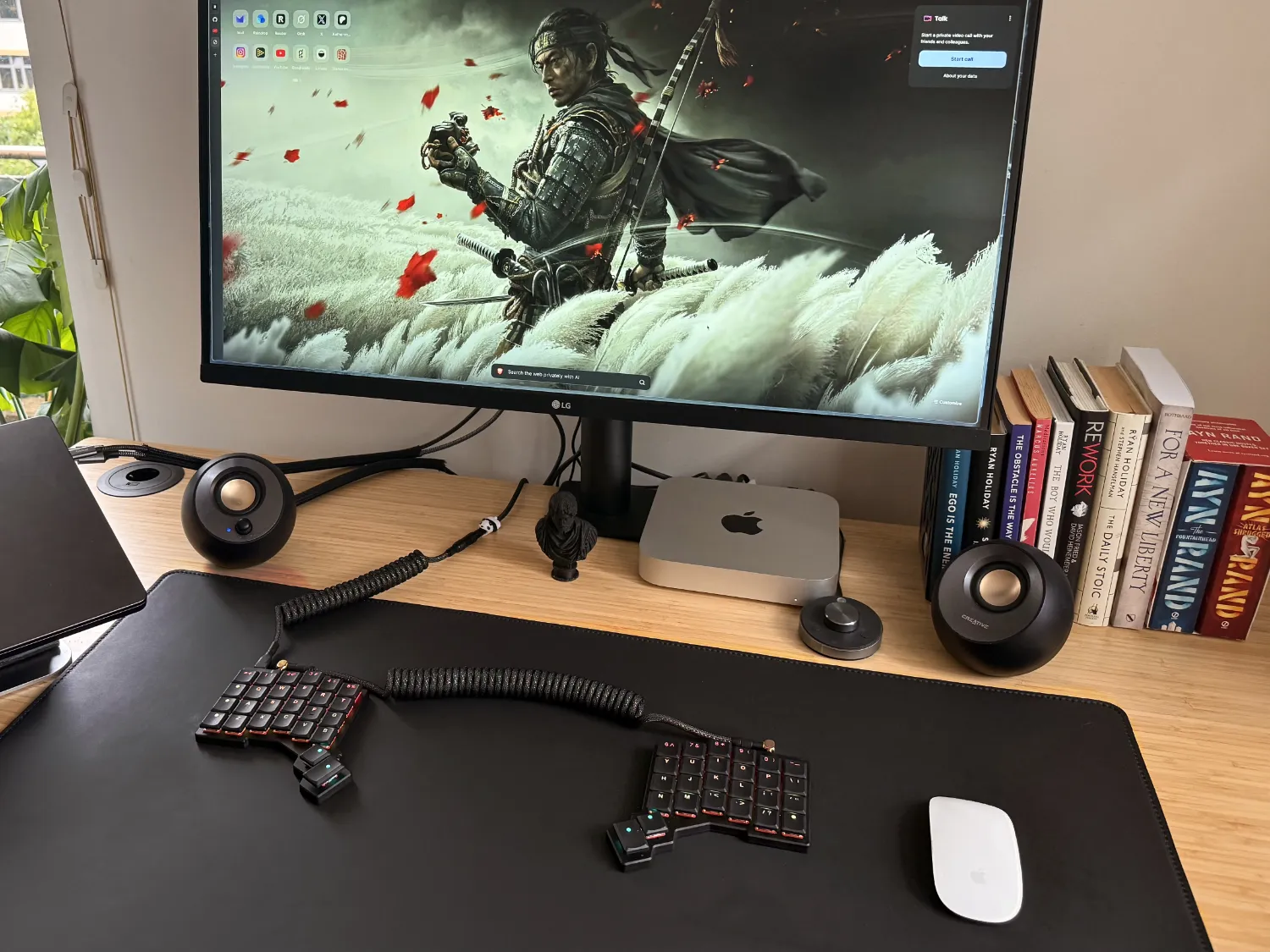 A modern desk setup with a large monitor displaying a samurai wallpaper, a split ergonomic keyboard, a Mac Mini, speakers, a white mouse, and a row of books on the right.
